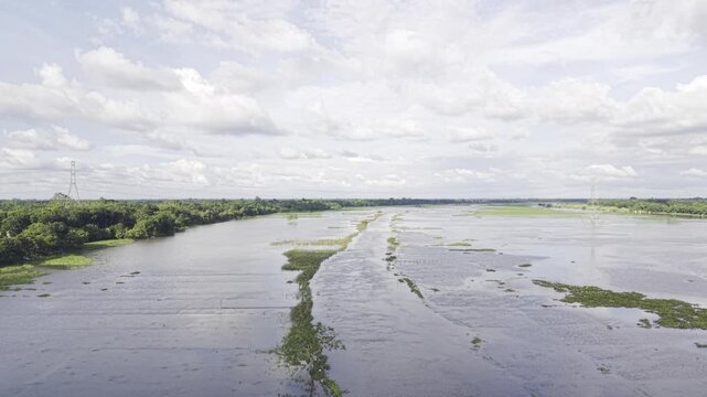 Drone moves forward over monsoon-flooded agricultural land in Kushtia, Bangladesh, revealing a narrow waterway, submerged fields, green grassy village banks, and a blue sky with white clouds.