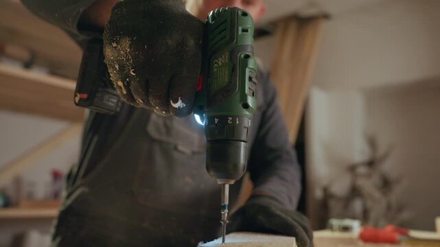 Man in workwear uses green cordless drill to make precise holes in wooden material. Sawdust scatters as drilling action progresses with workshop shelves and tools visible in soft indoor
