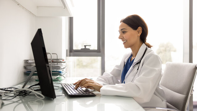 Young GP working on computer at desk in clinic office, reviewing patient medical records, writing prescriptions, updating patient information, check lab or test result, scheduling patient appointments