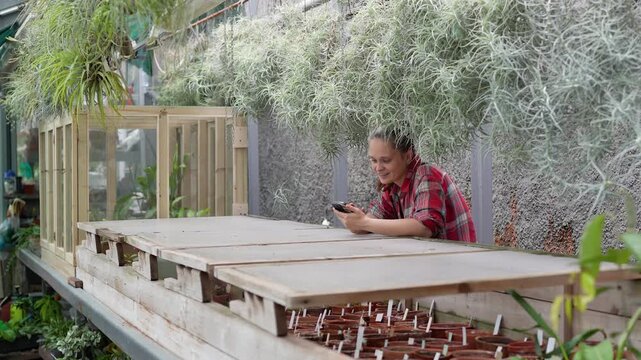 Cheerful woman botanist worker scrolling smartphone while standing under Tillandsia hanging plants, happy relaxed female gardener enjoying work break and browsing online on mobile in greenhouse.