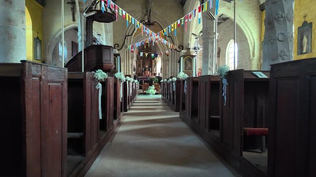 Slow motion forward movement through a decorated church nave with wooden pews, floral arrangements and colorful hanging ribbons in Veules-les-Roses, Normandy