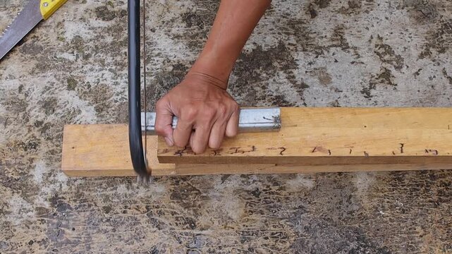 Close-up view of man sawing square metal tube placed on timber on concrete floor