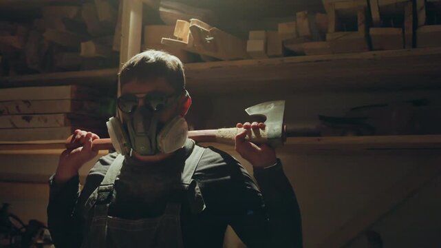 Man wearing safety equipment stands inside dimly lit workshop surrounded by wood. Man handles and carries axe indicating preparation for carpentry work