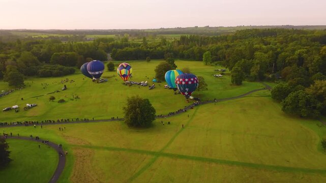 Families line paths through a wide green park as several colorful hot air balloons inflate and lift from the event field at dusk.