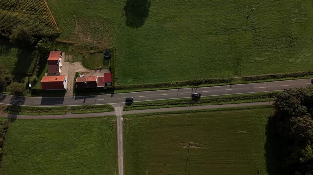 ars driving along rural road beside small farm property with red roof buildings, green fields, crossroad in America, harvested cornfield. Aerial drone top down flyover