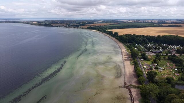 Curved sandy beach along Baltic Sea coastline with algae patterns in shallow water, campground, coastal road, dense trees, harvested fields in Germany, overcast sky. Aerial drone wide shot