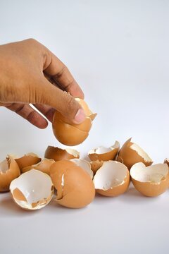 Studio shot of a human hand picking up a broken brown eggshell from a pile of scattered eggshells on a white background.