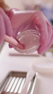 Vertical video. Beauty product. Alginate mask mixing. Unrecognizable cosmetologist woman hands in pink protective latex gloves using clear bowl for cosmetic procedure.