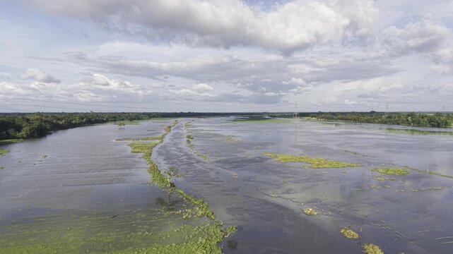 Drone ascends above monsoon-flooded agricultural land in Kushtia, Bangladesh, gradually revealing the extent of waterlogged fields, green village banks, a narrow waterway, and cloudy blue skies.