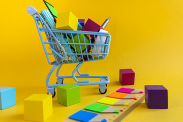 Shopping cart with cubes and stationery surrounded by scattered blocks on yellow background © Irina
