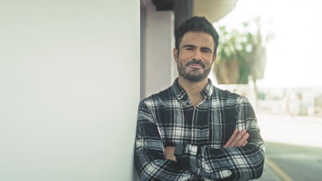 Man leaning against a building wall with arms crossed and smartwatch on wrist, wearing plaid shirt, outdoors by palm trees on street; calm reflection.