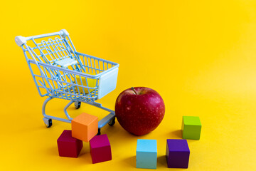 Empty shopping cart beside red apple and scattered colorful blocks on yellow background © Irina