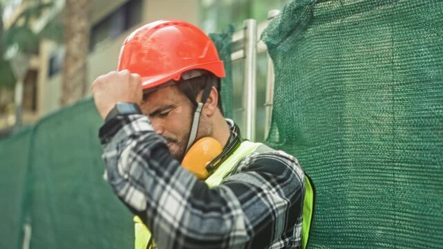 Man engineer adjusts hardhat then leans head against green fence at building with reflective vest and earmuffs; fatigue.