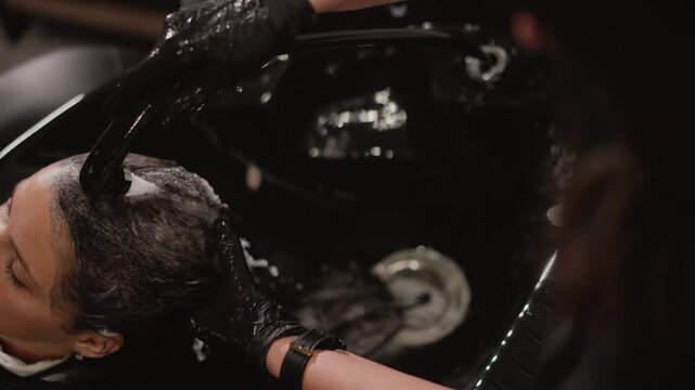 Close up view of client head as stylist applies treatment to wet hair. Stylist hands wearing black gloves and watch work over dark haired client positioned at salon sink