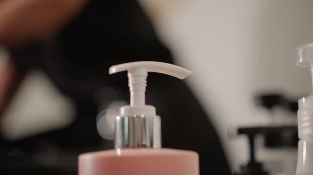 Close up shows beauty treatment pump bottle with blurred salon worker in background consistent focus on pink container and dispenser product care hair
