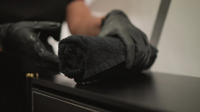 Gloved hand places rolled up dark fabric on smooth wood surface then removes it. Soft lighting highlights texture details in neutral background towel gloves