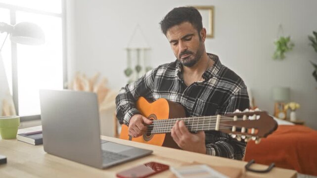 Man seated playing classical guitar, left hand on fretboard and right fingers strumming strings beside laptop on desk in studio; concentration.
