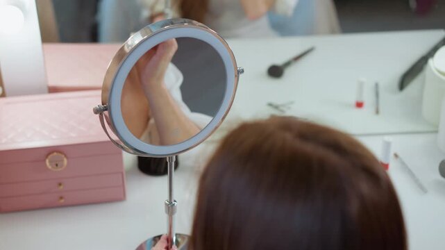 Shows round mirror with LED lighting reflecting part of scene. pink jewelry box and various makeup tools are visible on table beauty reflection cosmetics