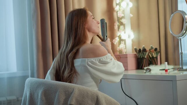 Woman straightens her hair in front of vanity mirror surrounded by makeup brushes and beauty products. Woman is seated at white dressing table with soft lights near window curtains