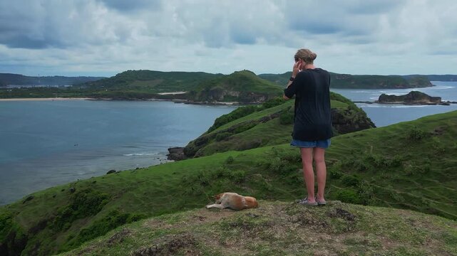 Caucasian woman and dog overlooking coastline while capturing photo with phone, standing on grassy promontory under cloudy sky, expansive seascape, attentive stance, pet resting beside, Lombok
