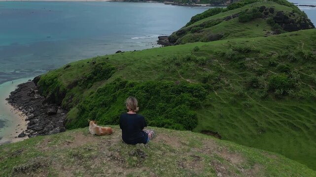 Caucasian woman and dog sitting on cliff overlooking turquoise bay, panoramic aerial view of clear water, sandy cove, green slopes, tranquil mood, gentle breeze, Bukit Merese, Lombok, Tanjung Ann