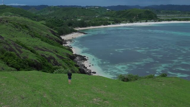 Woman walking hill above turquoise bay, solo hiker pauses to admire coastal vista, green slopes, sandy beach, clear shallow water, distant mountain ridge, Bukit Merese, Lombok, Tanjung Ann