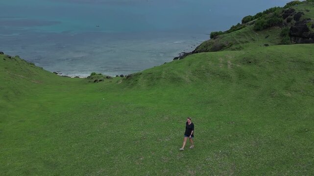 Woman walking hill above turquoise bay, solo hiker pauses to admire coastal vista, green slopes, sandy beach, clear shallow water, distant mountain ridge, Bukit Merese, Lombok, Tanjung Ann