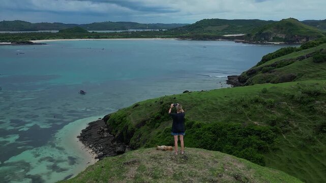 Woman With Dog Taking Selfie On Grassy Cliff Overlooking Turquoise Ocean And Small Boat, Casual Outfit And Sunglasses, Joyful Travel Vibe, Dramatic Shoreline And Panoramic Aerial View,Lombok