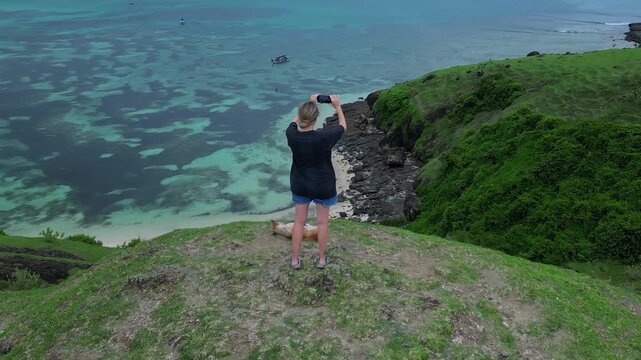 Woman With Dog Taking Selfie On Grassy Cliff Overlooking Turquoise Ocean And Small Boat, Casual Outfit And Sunglasses, Joyful Travel Vibe, Dramatic Shoreline And Panoramic Aerial View,Lombok