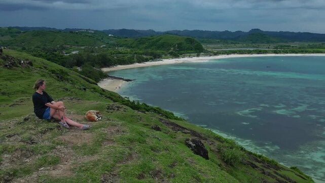 Caucasian woman and dog sitting on cliff overlooking turquoise bay, panoramic aerial view of clear water, sandy cove, green slopes, tranquil mood, gentle breeze, Bukit Merese, Lombok, Tanjung Ann