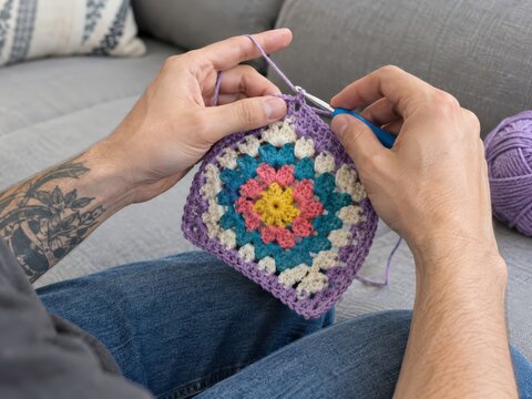 Young man crochets a colorful granny square on a sofa