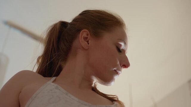 Close up portrait captures side profile of young lady with red hair tied in low ponytail. Woman wears white camisole and soft natural lighting illuminates her skin against neutral indoor