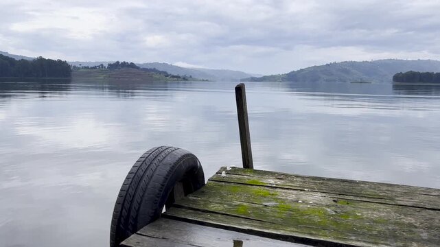 Calm water reflects cloudy skies on Lake Mutanda, southwestern Uganda, with a weathered wooden dock, tire fender and forested hills surrounding the volcanic highland lake, static wide view.