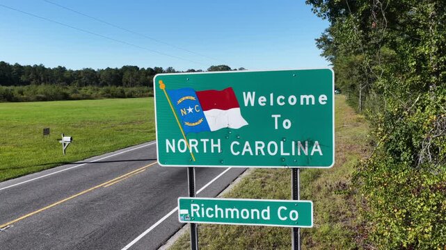 Sunlight falls evenly across a Welcome to North Carolina Richmond County sign and a hay strewn field, the two lane road running empty toward the tree line. Aerial orbit shot