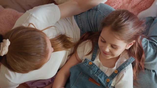 Woman gently touches hair of young girl while lying together on soft pink fabric emphasizing maternal affection. intimate moment is set in cozy domestic environment with warm light
