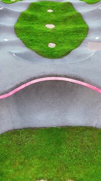 Oblique aerial view of a modern asphalt pump track with rolling humps, pink guide lines and green islands featuring a heart-shaped stone landscaping detail.
