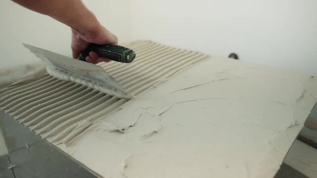 Close-up of a worker combing fresh tile adhesive with a notched trowel, creating parallel ridges across the floor substrate before laying the next tile.