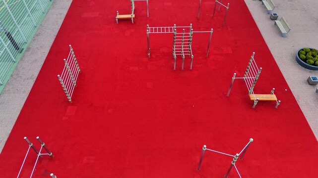 Oblique drone view of an empty outdoor calisthenics park with monkey bars, climbing ladders and bodyweight stations set on vivid red rubber flooring.