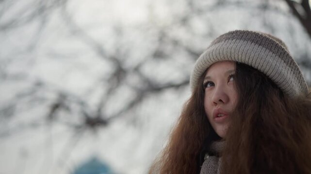 Woman stands outside on cold day. Woman wears knitted beanie with fur trim over her ears and matching scarf while background shows bare trees outdoor winter