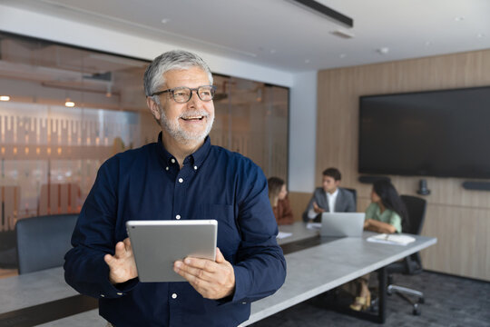 Smiling senior businessman holding digital tablet, communicating, consulting online, standing in conference room at group office meeting. Project management, corporate workflow, business, copyspace