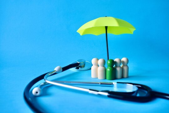 Wooden peg figures standing under a yellow umbrella next to a medical stethoscope on a blue background, symbolizing health insurance, medical coverage, and family protection.