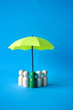 Wooden peg figures with a green leader figure standing under a small yellow umbrella against a vibrant blue background, representing leadership, team protection, and group insurance concepts.
