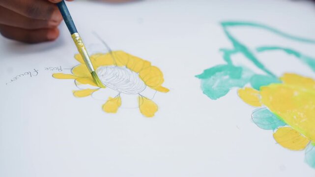 Close up shows steady focused process where hands carefully apply bright colors to pre drawn outlines. hand holding brush fills in details of yellow petals and centers on white paper