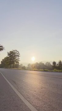 Vertical footage of a quiet rural road at sunset, with warm sunlight reflecting on the asphalt, trees, hazy mountains and a peaceful countryside atmosphere.