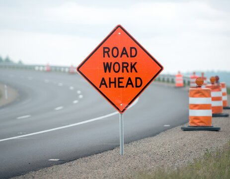 Orange diamond "ROAD WORK AHEAD" sign beside a highway with orange cones and barrels marking a road construction zone on wet asphalt.