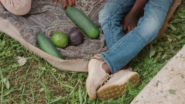 Children sit on grass legs visible as they interact with fresh produce including cucumber avocado and lime placed near them. Patterned cloth serves as backdrop for scene