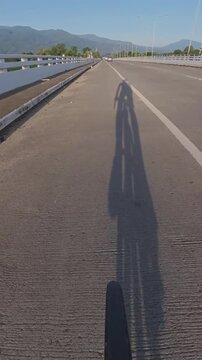 Vertical cycling POV riding across a bridge, with a long cyclist shadow on the road, distant mountains and scenic surroundings under clear morning light.