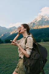 Naklejka premium A young woman with a backpack stands in a mountain meadow covering her face with one hand while holding a phone, enjoying a moment of relaxation in nature. Woman lifestyle concept.