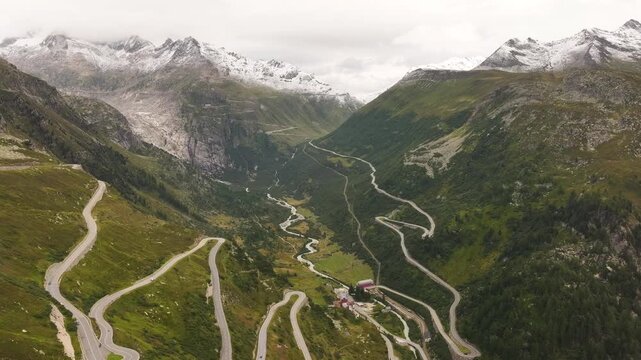 Aerial view across winding alpine roads and hairpin bends near Gletsch, Switzerland, with a river, small settlement and rugged snowy peaks framing the valley.