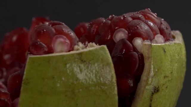 Spinning pomegranate halves reveal glossy ruby red seeds and pale inner membranes against a dark background, highlighting the green rind and juicy fruit texture in macro detail.